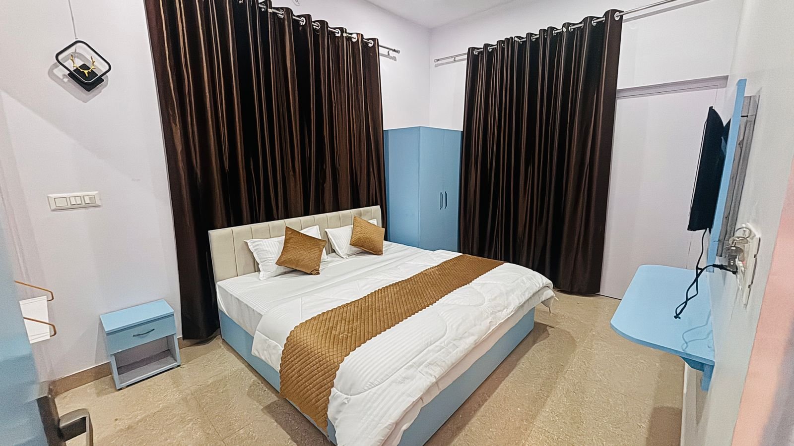 Modern guest bedroom with white linens, brown runner, light blue wardrobe, TV, and dark floor-to-ceiling curtains